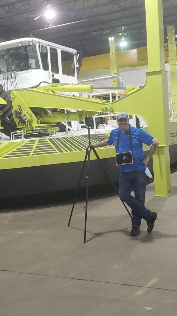 A technician in a blue shirt standing next to a scanning tripod in a warehouse with a large industrial vehicle in the background.